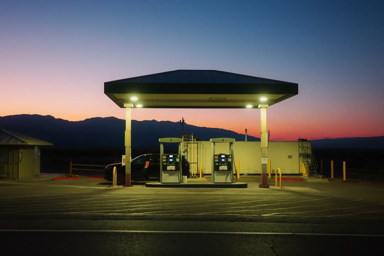 An illuminated gas station against mountain silhouettes and a pink sunset sky